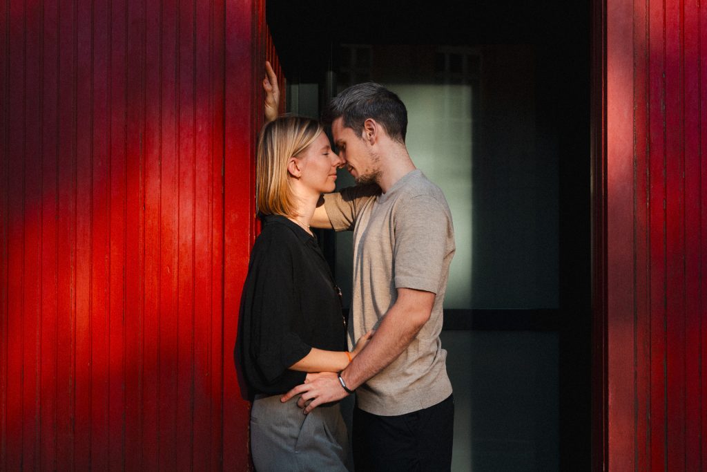 Couple qui s'embrasse lors d'une séance engagement. Il est placé dans l'encadrement d'une porte et les murs rouges sont de part et d'autre