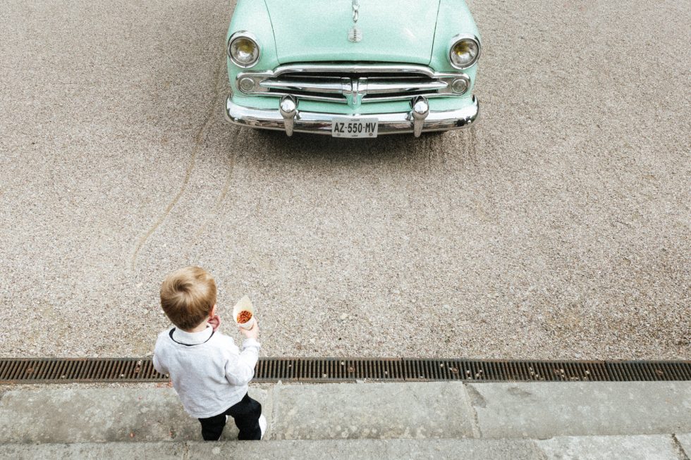 Un garçon regarde une voiture verte d'où vont sortir des mariées, il a un cornet avec des pétales de fleurs dans la main