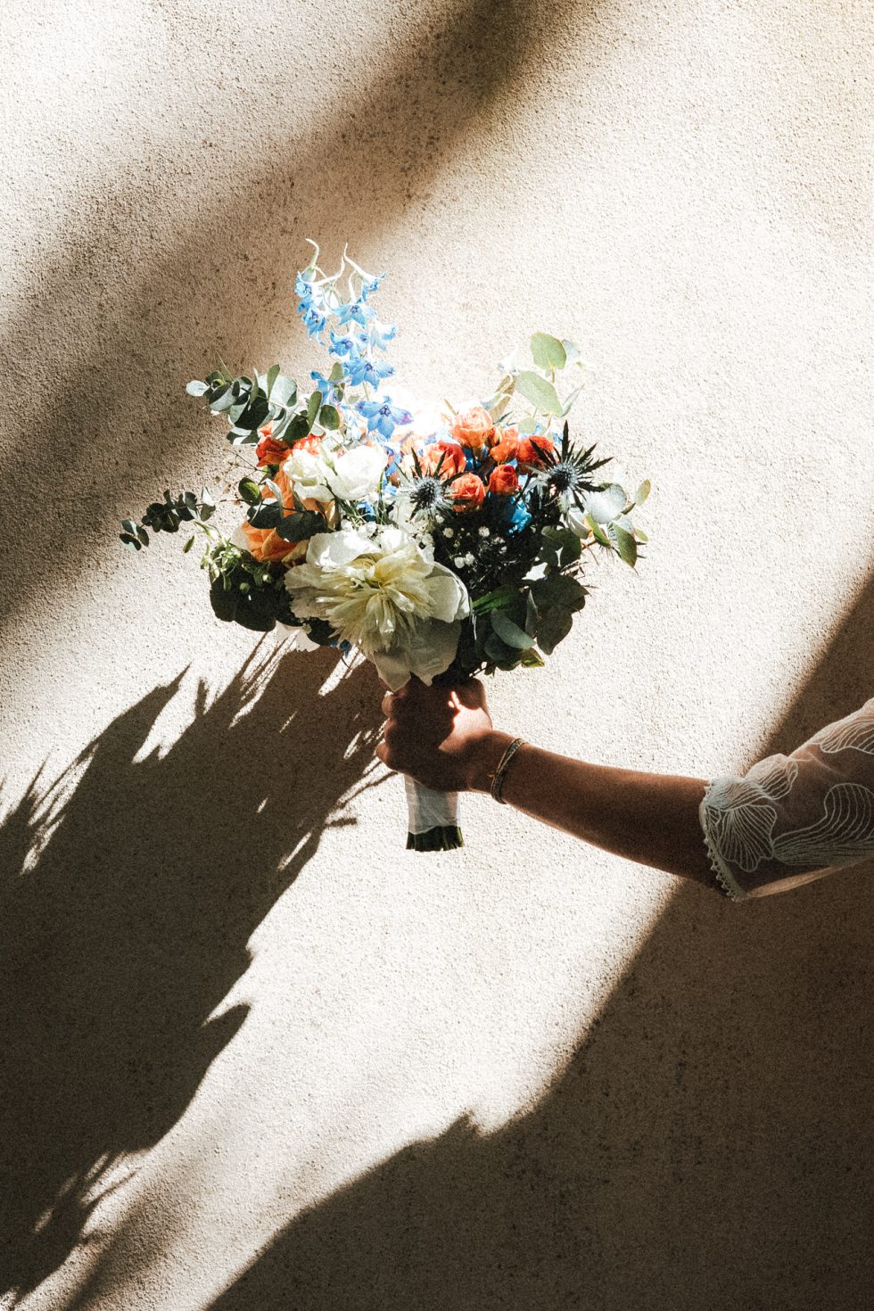 La mariée tient son bouquet de fleurs contre le mur. On ne voit que son bras et il y a un jeu d'ombres et lumières
