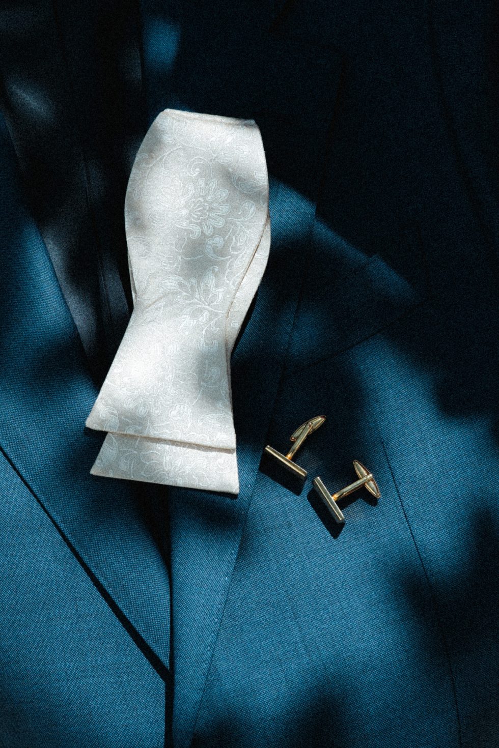 Détail du costume du marié, nœud papillon ivoire et boutons de manchette dorés baignés de lumière