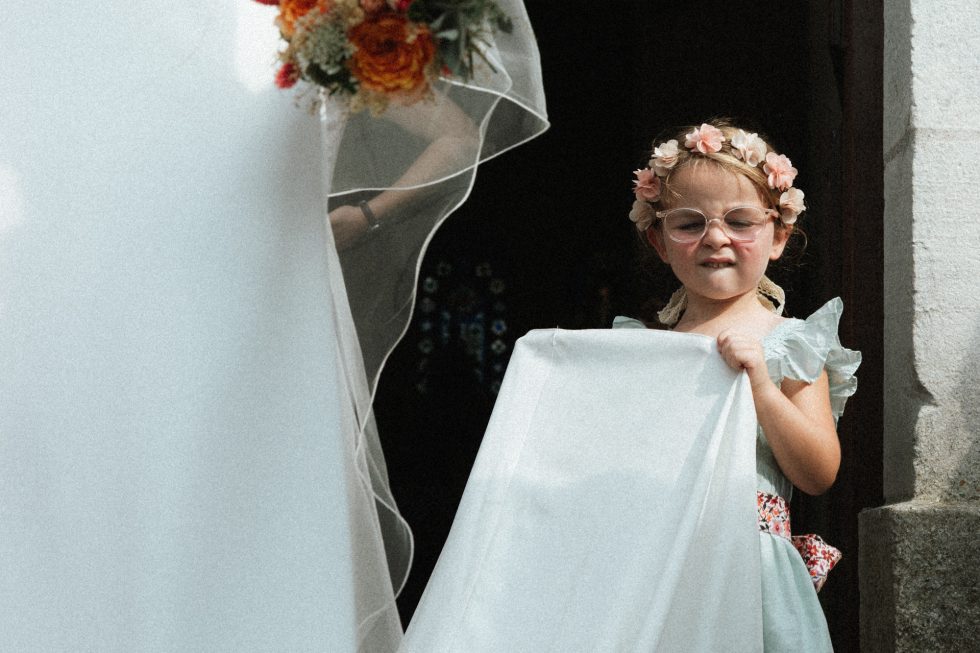 Une demoiselle d'honneur tient la traine de la mariée à la sortie de l'église. Elle a une mimique amusante.
