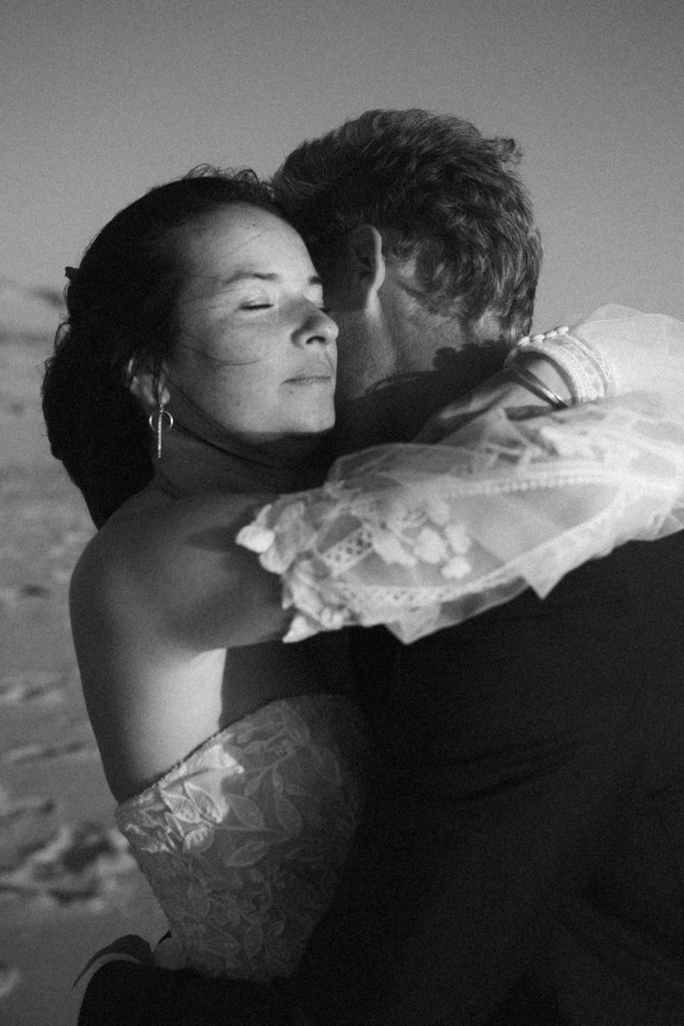 Séance jeunes mariés sur la plage de Lacanau Océan . La mariée enlace délicatement son mari, son visage est baigné par la lumière douce du soleil.