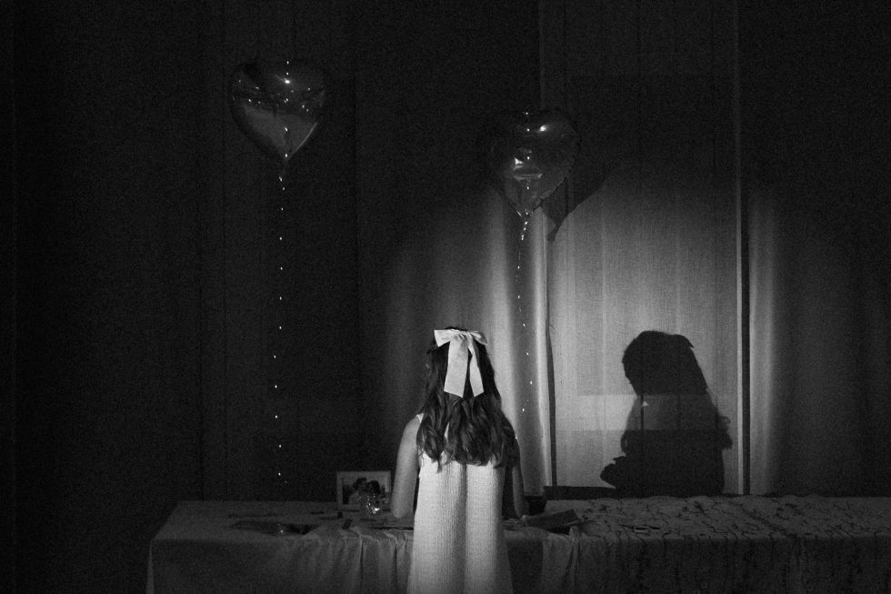 Petite fille de dos, éclairée par une lumière douce, écrivant un mot sur le livre d’or entre deux ballons en forme de cœur.