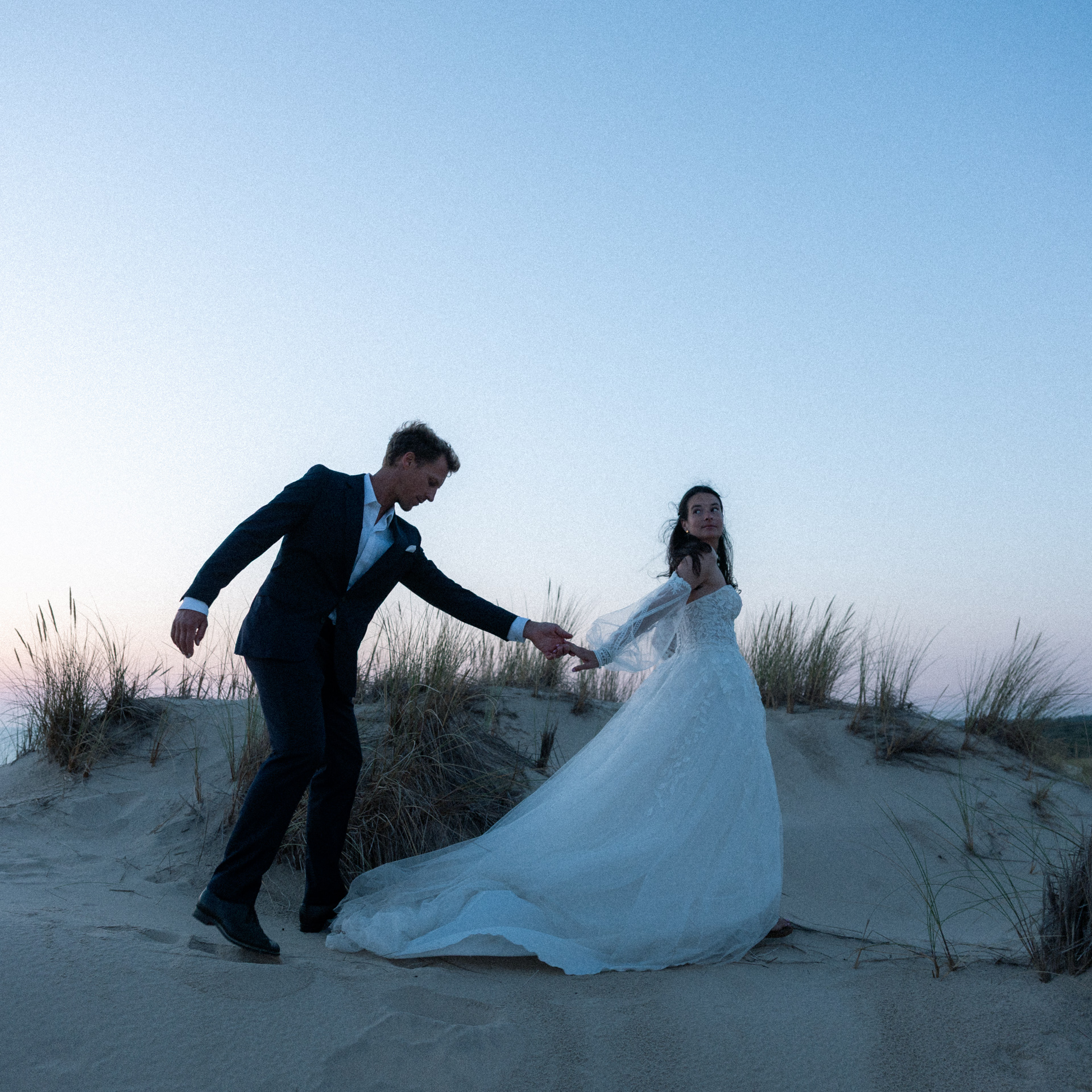 Séance jeunes mariés dans les dunes à Lacanau Océan. La mariés tient son mari par la main, elle le regarde