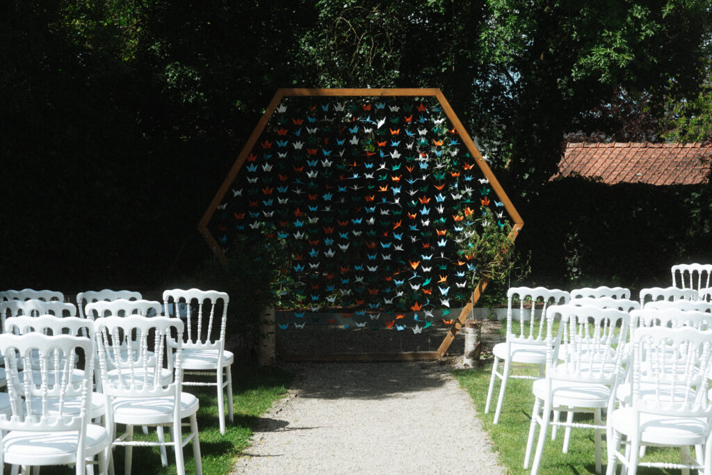 Allée de cérémonie laïque décorée de chaises blanches et d’une structure ornée de guirlandes de grues en papier dans un cadre naturel