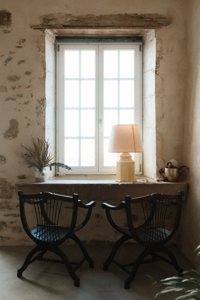Fenêtre ancienne avec lampe et fauteuils dans un salon rustique du Château Ferme d’Aboncourt, ambiance douce et chaleureuse.