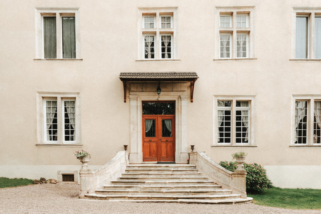 Façade du Château de Morey avec escalier en pierre et porte en bois, entourée d’un jardin soigné"