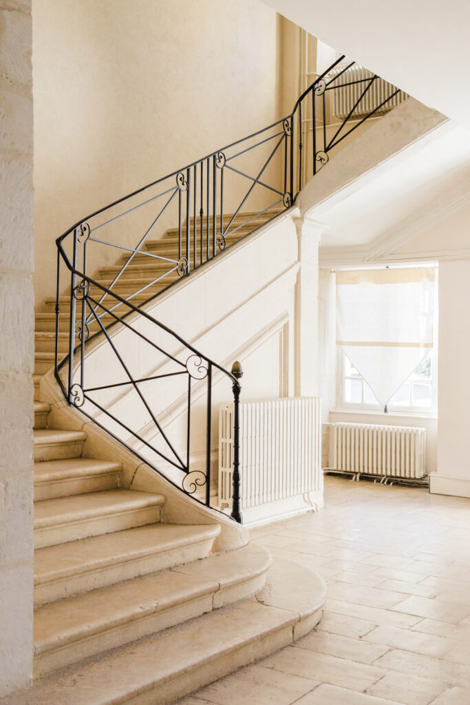 Escalier intérieur en pierre avec rampe en fer forgé dans le Château de Vandeleville, baigné de lumière naturelle