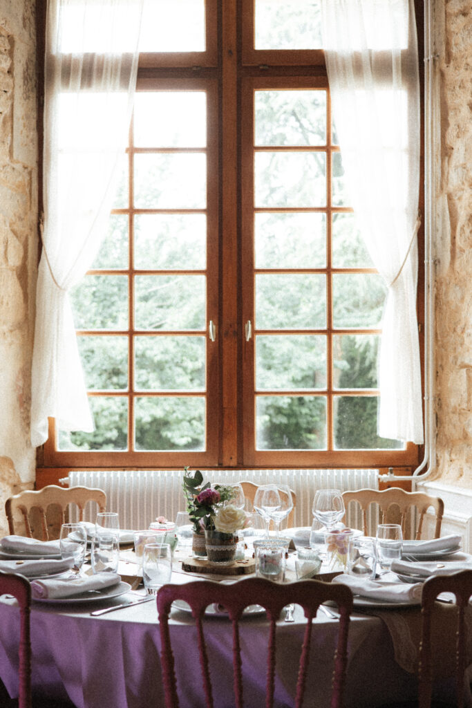 Table de mariage dressée devant une grande fenêtre avec un voilage, vaisselle élégante et centre floral au Château de Morey