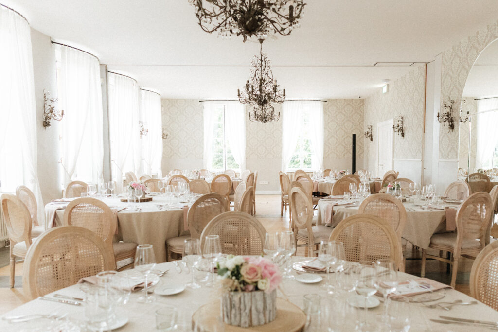 Salle de réception classique et élégante avec tables rondes dressées, chaises en bois clair et lustres au Château de Schengen au Luxembourg