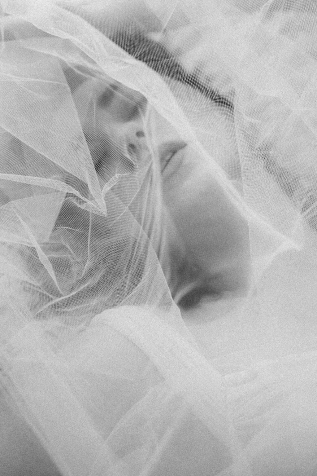 Gros plan flou et délicat sur une mariée en lingerie blanche, enveloppée dans un voile transparent pendant une séance boudoir de la mariée