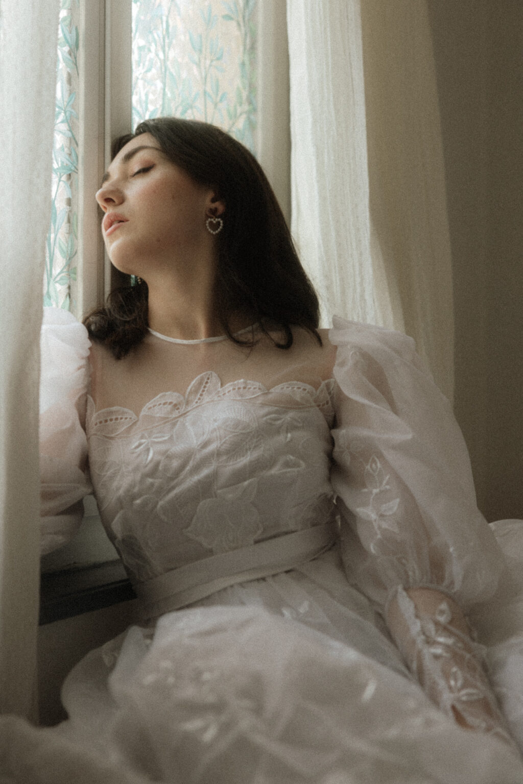 Portrait doux d’une mariée en robe blanche brodée près d’une fenêtre, avec boucles d’oreilles cœur et lumière naturelle pendant une séance boudoir de la mariée.