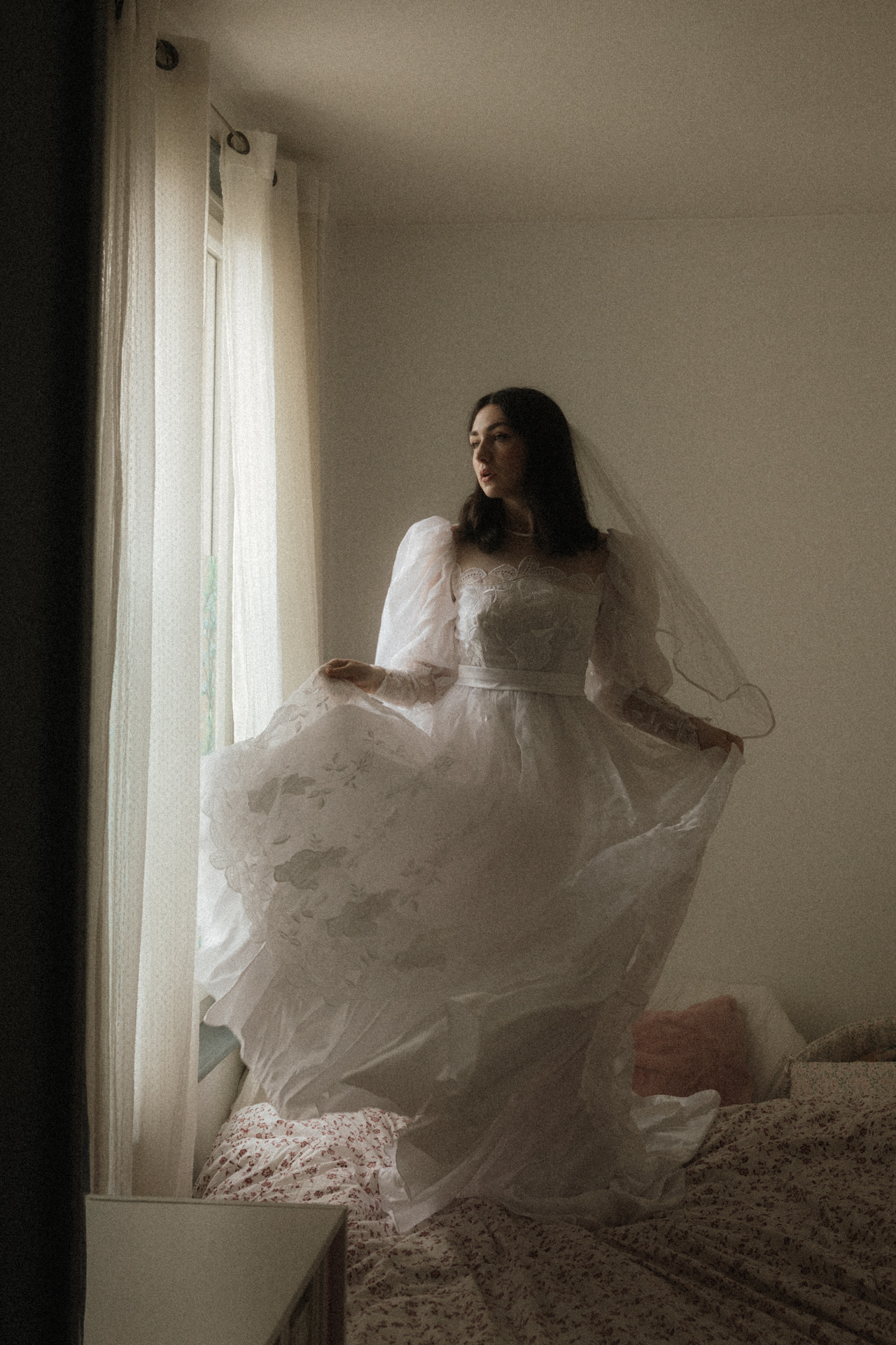 Mariée en robe blanche brodée posant près d’une fenêtre, entourée de voile et de lumière naturelle.