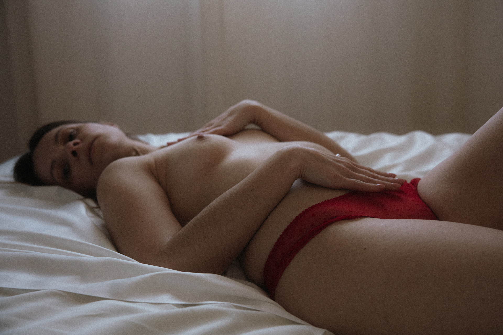 Séance boudoir en Lorraine avec lingerie rouge, femme allongée sur un lit dans une lumière naturelle tamisée.