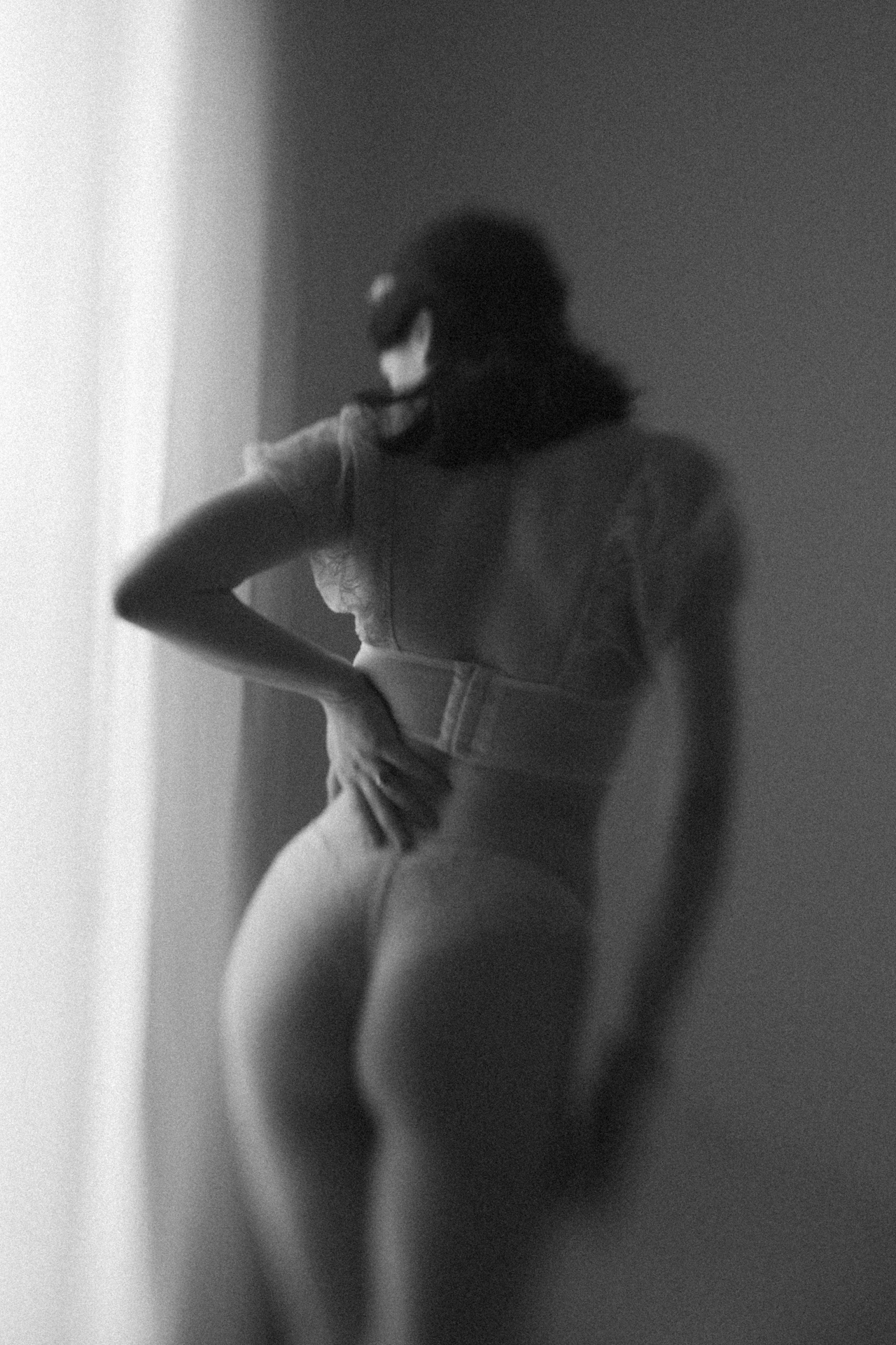 Mariée de dos en lingerie blanche devant une fenêtre, dans une photographie noir et blanc au rendu vaporeux. Séance boudoir de la mariée.