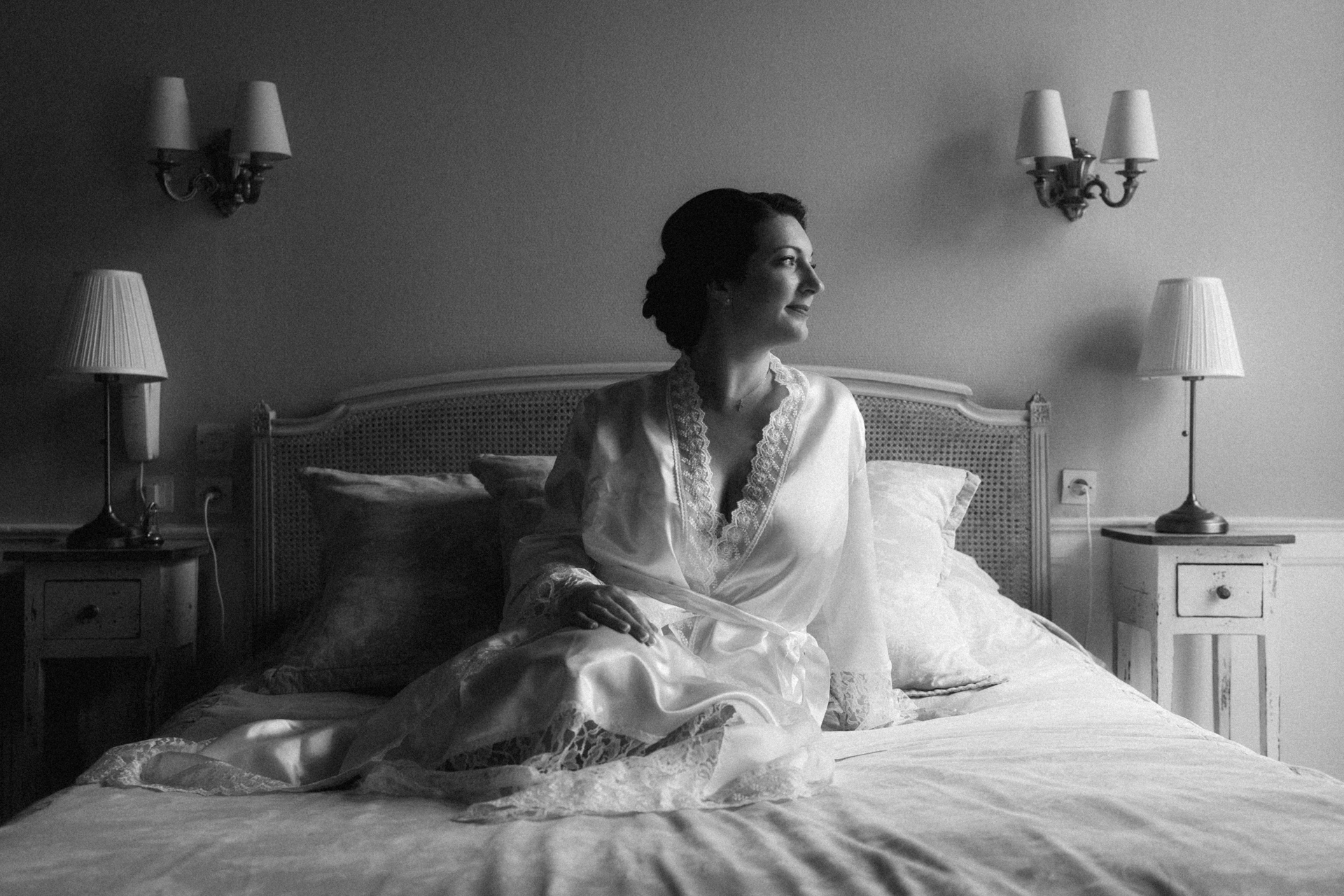 Séance boudoir de la mariée. La mariée, vêtue d'un déshabillé en satin blanc est assise sur le lit et regarde vers la lumière, dans une ambiance élégante