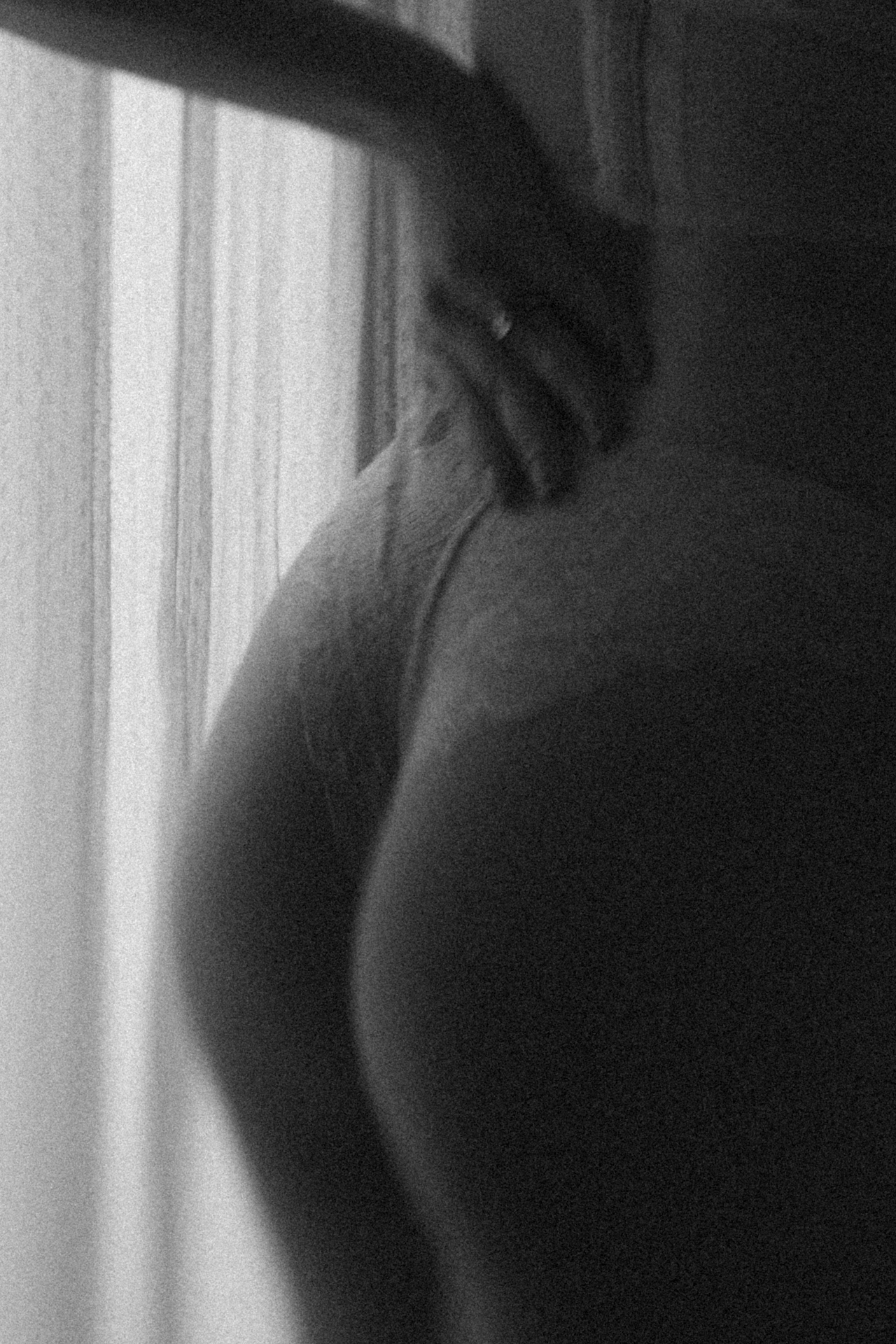 Détail d'une mariée de dos en lingerie blanche devant une fenêtre, dans une photographie noir et blanc au rendu vaporeux. Séance boudoir de la mariée.