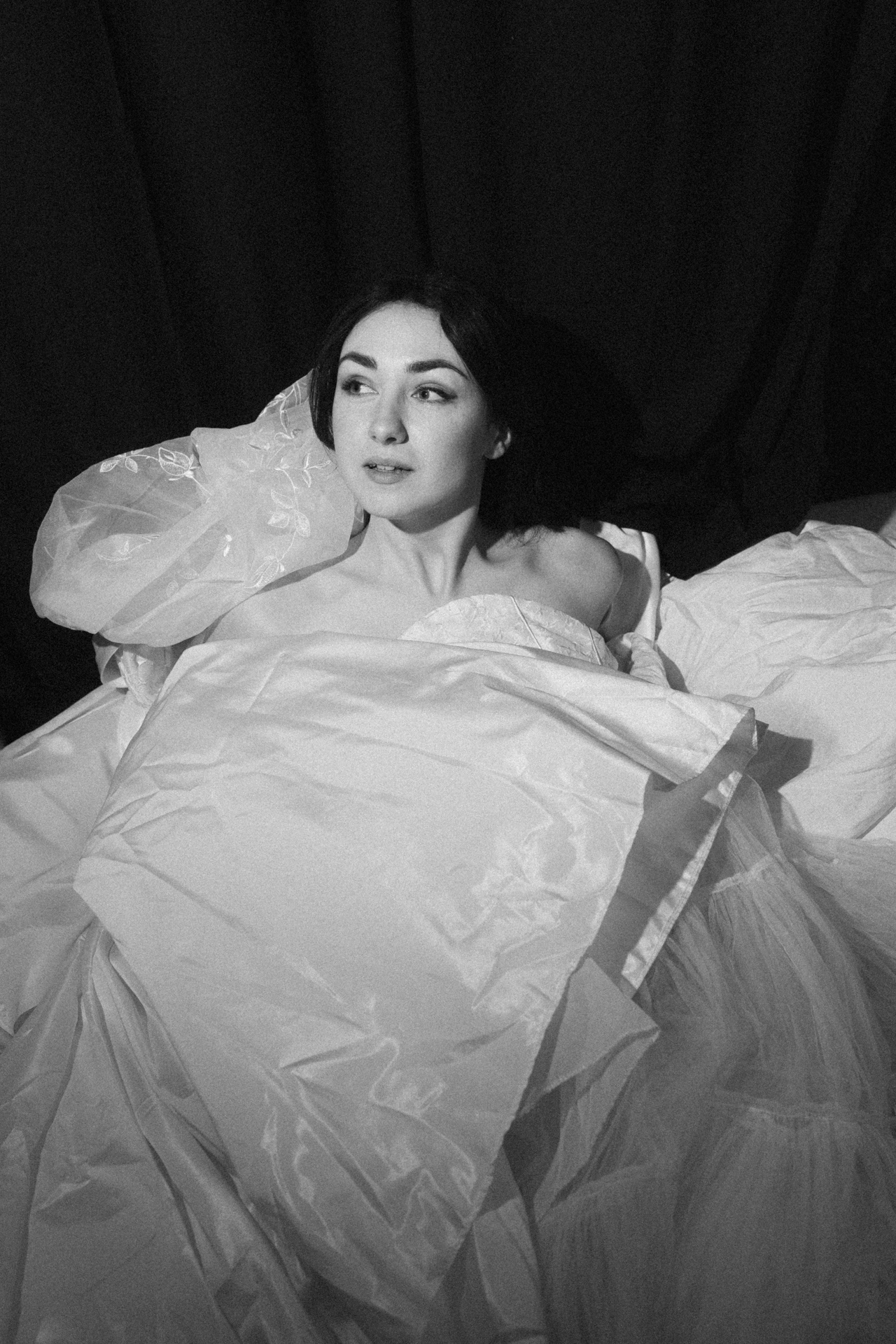 Portrait noir et blanc d’une mariée allongée au milieu de ses robes dans une ambiance éditoriale. Pendant une séance boudoir de la mariée